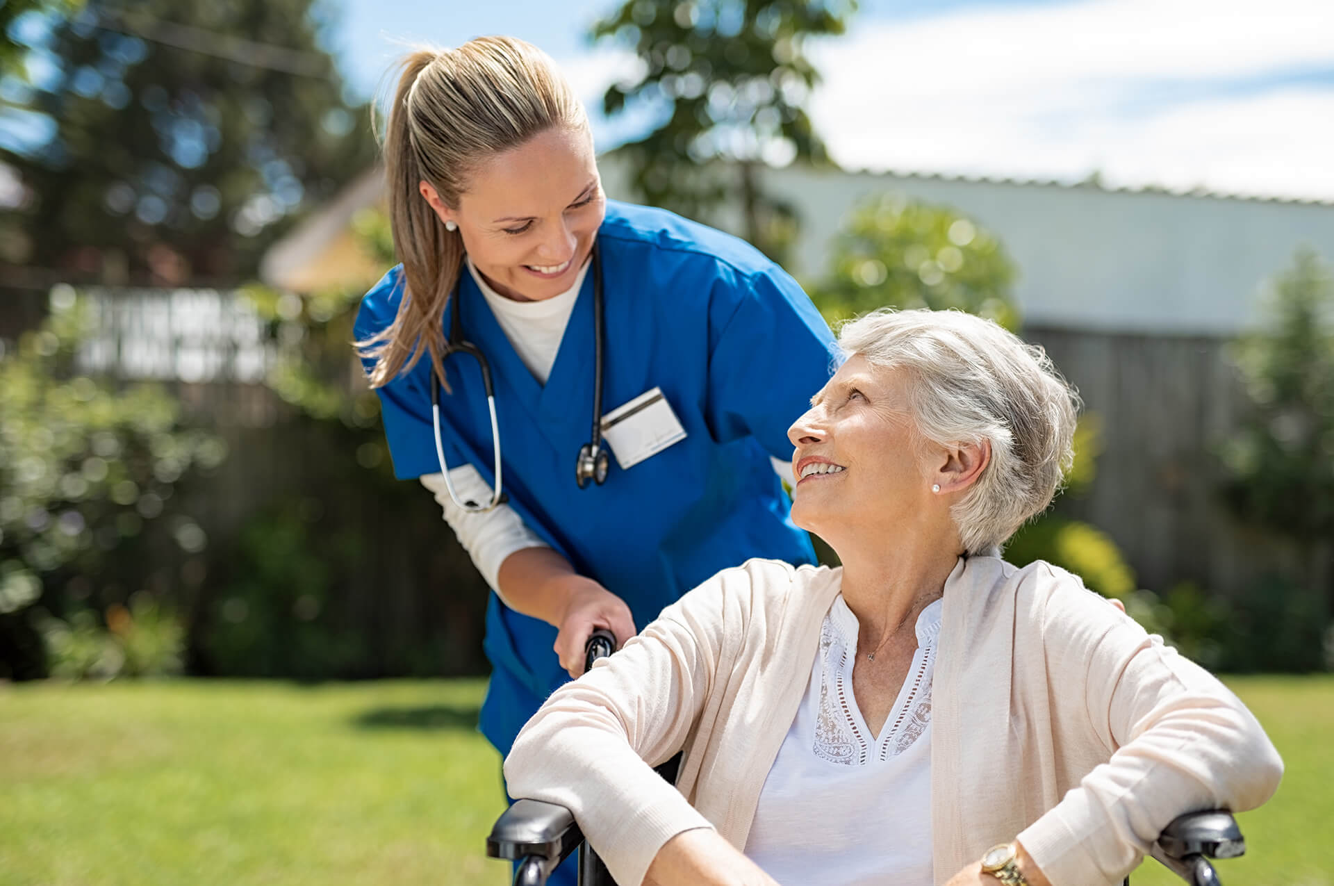Smiling nurse providing care to an elderly woman in a wheelchair outdoors, representing Sunshine Home Care Services’ compassionate and professional senior care.